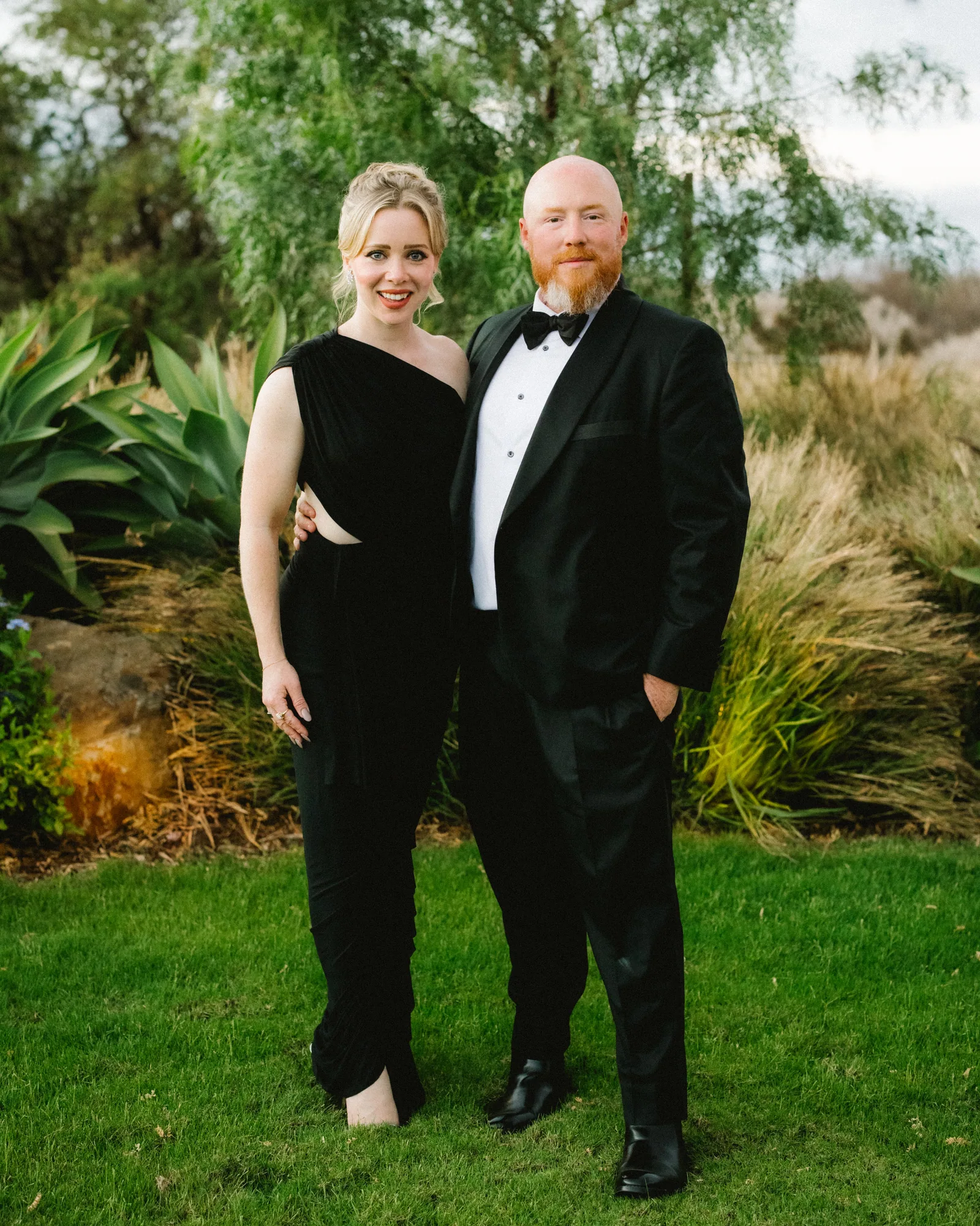 A couple in black tie photographed on a lawn against tropical foliage
