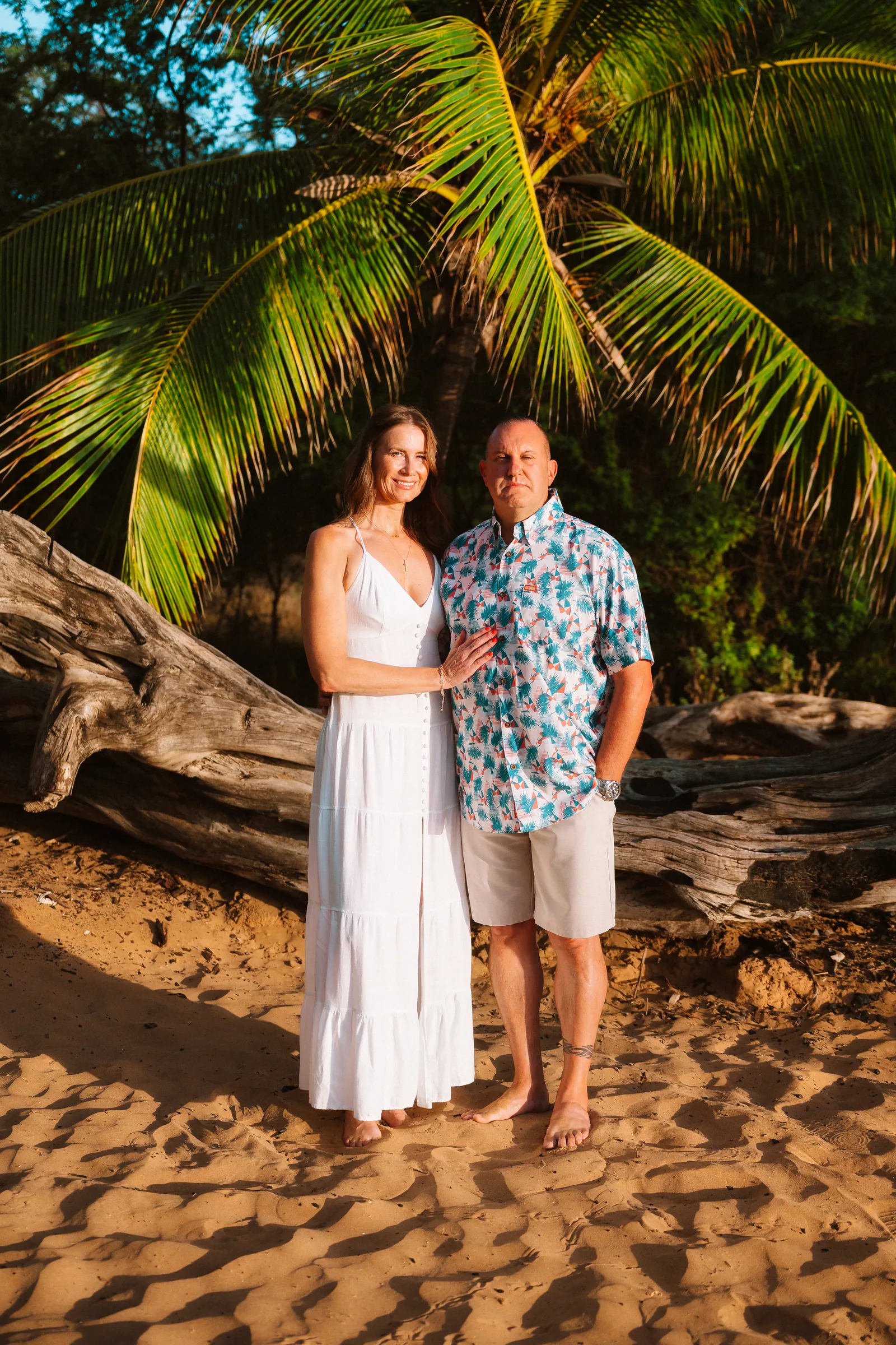A couple standing beside driftwood under palms on the beach