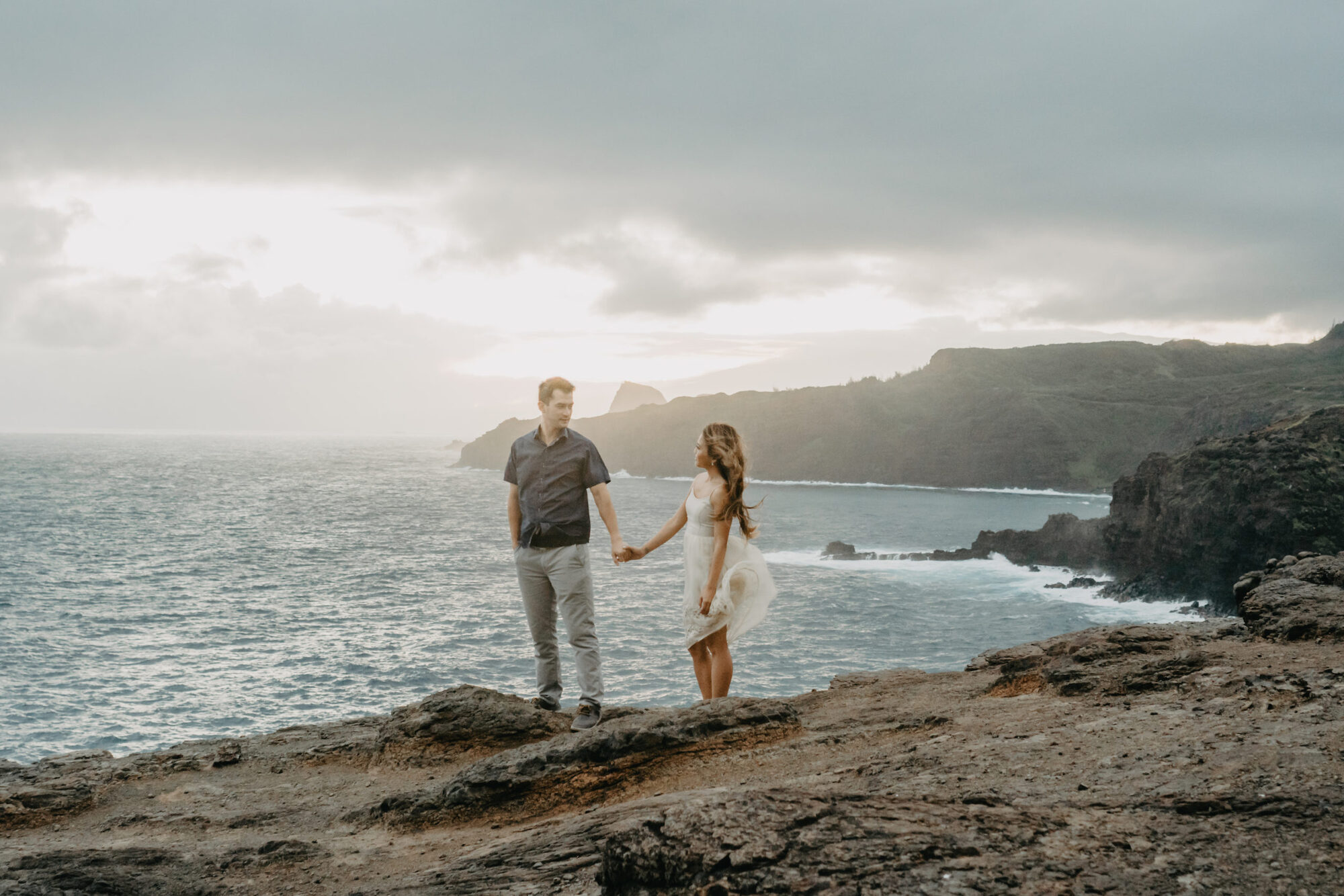 A couple holding hands on a rocky Maui sea cliff overlooking the ocean at dusk during a session with Jessie Hawaii Photography