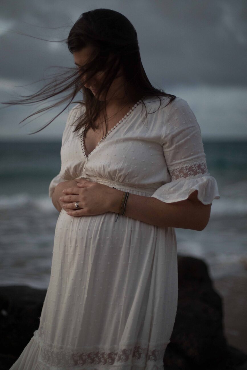Jessie Hawaii Photography, Pregnant woman in white dress on beach, gently holding her belly.