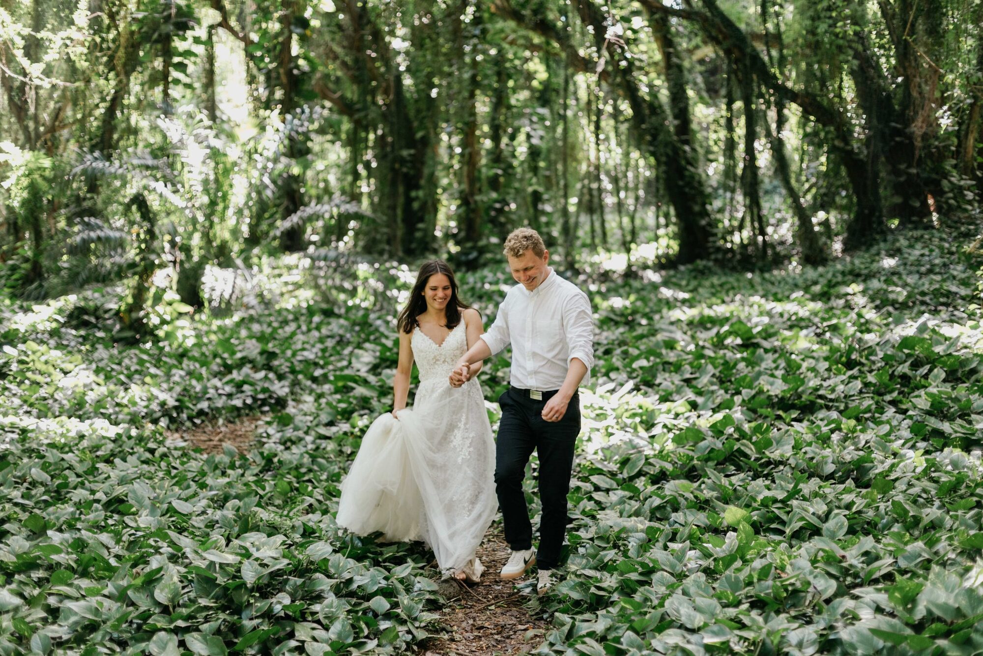 Jessie Hawaii Photography, Happy bride and groom holding hands, walking through a lush green forest.
