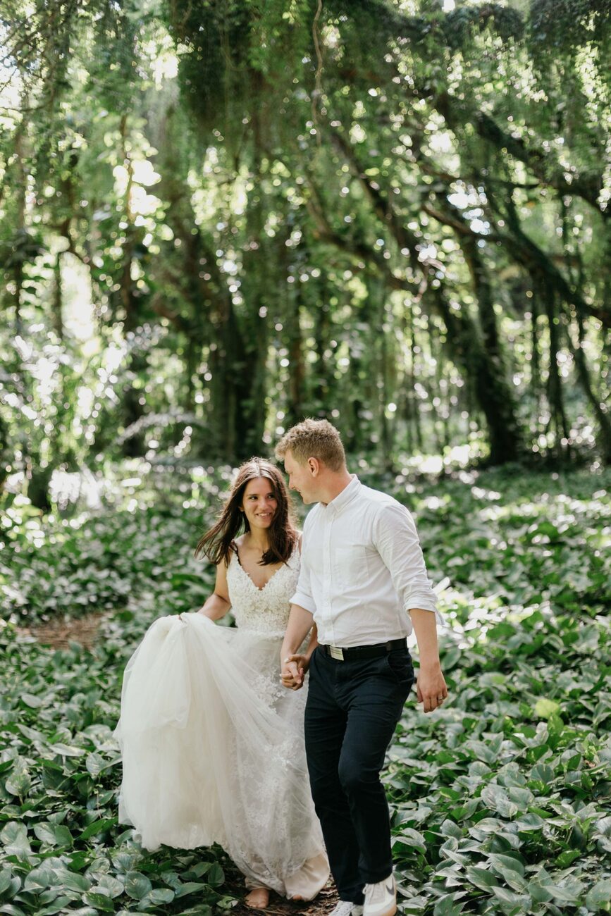 Jessie Hawaii Photography, Happy bride and groom holding hands, walking barefoot through a lush green forest.