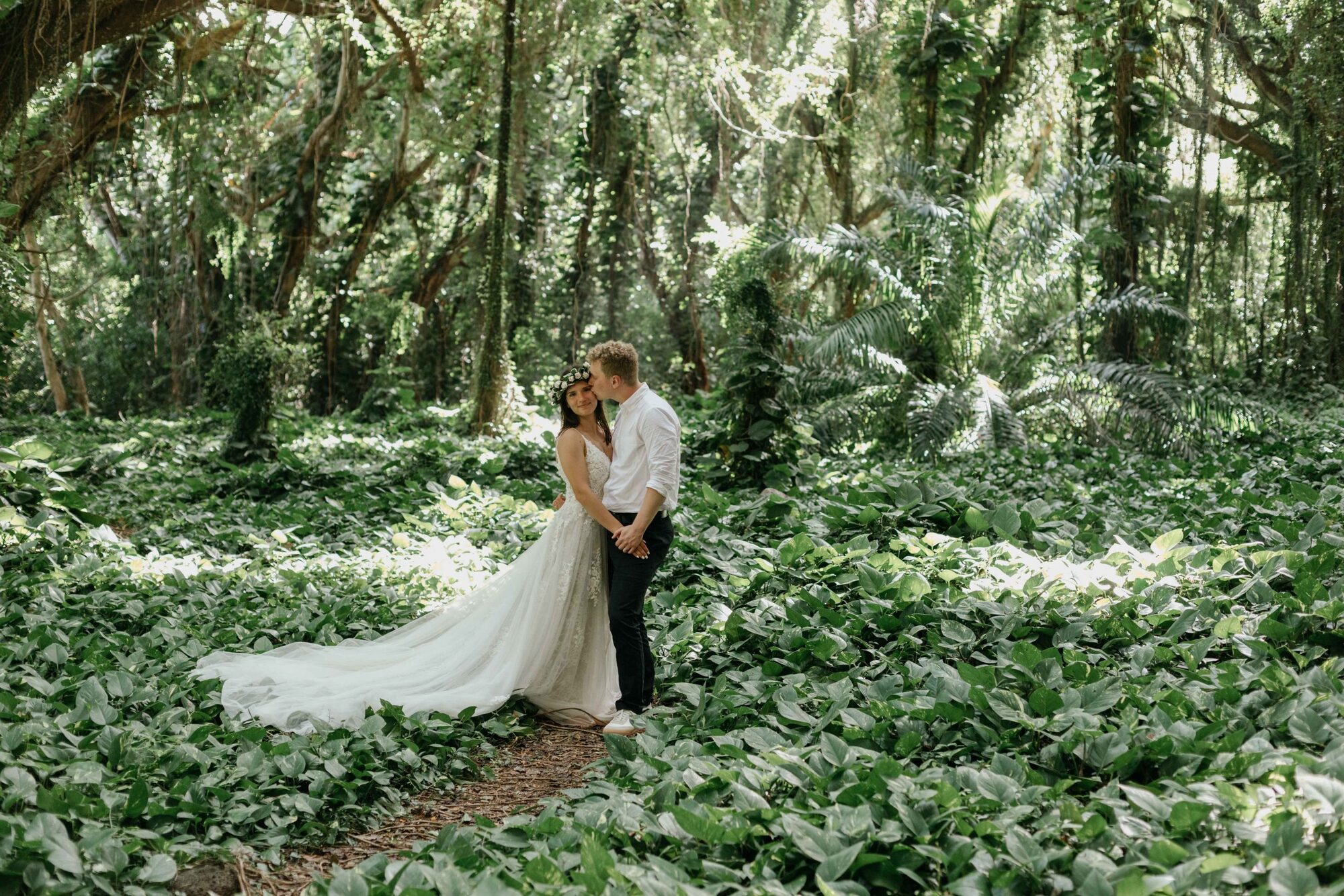 Jessie Hawaii Photography, Bride and groom embrace in lush green forest, wedding dress trailing on ground.